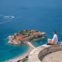 Mężczyzna siedzi na murze, patrząc na wyspę Sveti Stefan. Czy Czarnogóra jest w Unii? Piękny widok na Adriatyk.