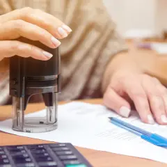 Dłoń z pieczątką nad dokumentem, obok długopis i kalkulator. To dowód tymczasowy, który zaraz zostanie zatwierdzony.