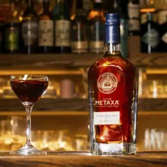 Butelka greckiego alkoholu Metaxa 12 gwiazdek i kieliszek z ciemnym trunkiem na tle baru.