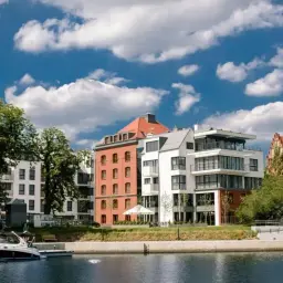 Nowoczesne budynki nad wodą, przypominające luksusowy almond hotel, z łodzią przy nabrzeżu i błękitnym niebem.