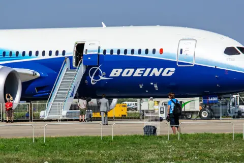 Ogromny samolot Dreamliner Boeing z otwartymi drzwiami i schodkami. W tle ludzie i sprzęt lotniskowy.