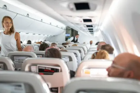 Kobieta w białej sukience stoi w przejściu samolotu, podczas gdy pasażerowie siedzą w rzędach. Widać lekkie turbulencje w samolocie.