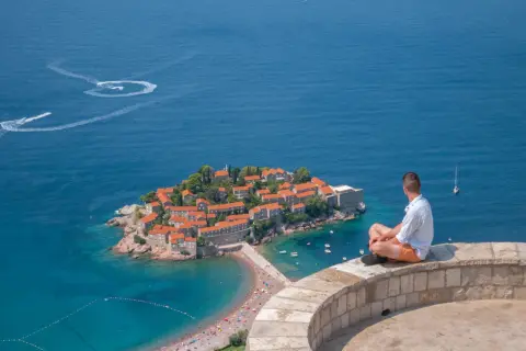 Mężczyzna siedzi na murze, patrząc na wyspę Sveti Stefan. Czy Czarnogóra jest w Unii? Piękny widok na Adriatyk.