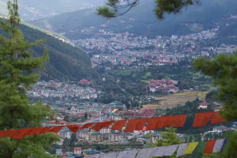 Widok na stolicę Bhutanu, Thimphu, z góry. W oddali widać gęsto zabudowane wzgórza, a na pierwszym planie wiszą kolorowe flagi modlitewne.