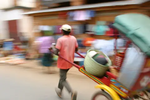 Madagaskar ludność w ruchu. Mężczyzna ciągnie wózek z koszem i towarami, pośród zamazanych postaci na targu.