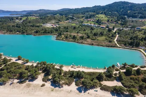 Turkusowa laguna na Kassandrze, otoczona zielonymi wzgórzami i sosnowym lasem. W oddali widać zabudowania i plażę z łodziami.