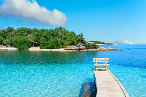 Albania - wczasy samolotem all inclusive. Drewniane molo z ławką prowadzi do krystalicznie czystej wody. Zielona wyspa w tle.