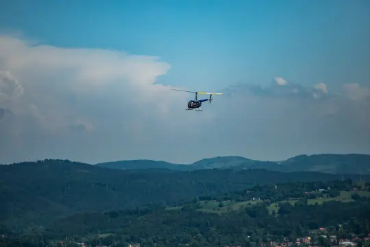 Niebieski helikopter wykonuje lot nad zielonymi wzgórzami i lasami.