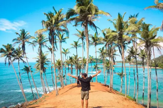 Wspaniałe loty na Sri Lankę! Człowiek z rozłożonymi rękami na tle palm i turkusowego oceanu.