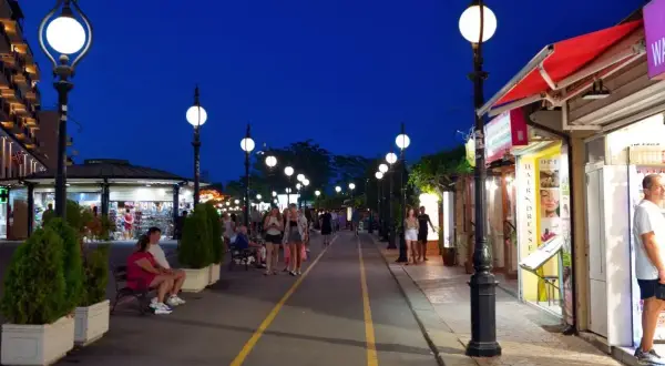 Wieczorna promenada w Złotych Piaskach tętni życiem. Ludzie spacerują, sklepy kuszą światłem, a latarnie oświetlają drogę.