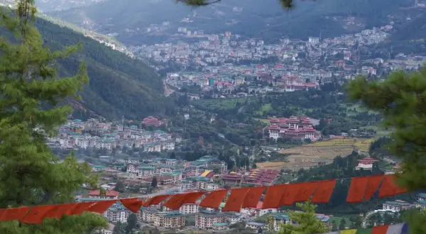 Widok na stolicę Bhutanu, Thimphu, z góry. W oddali widać gęsto zabudowane wzgórza, a na pierwszym planie wiszą kolorowe flagi modlitewne.