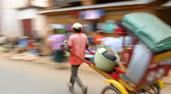 Madagaskar ludność w ruchu. Mężczyzna ciągnie wózek z koszem i towarami, pośród zamazanych postaci na targu.