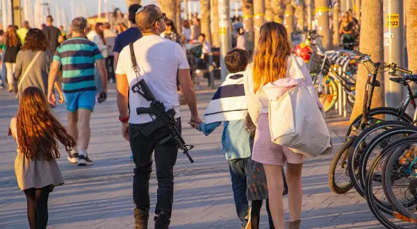 Rodzina spaceruje po promenadzie, mężczyzna ma przy sobie broń. W tle widać rowery i palmy, co może sugerować, że to Izrael. Liczba ludności Izraela jest wysoka, a bezpieczeństwo jest priorytetem.