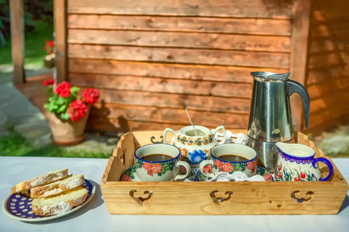 Drewniana taca z kawą, mlecznikiem, cukiernicą i ciastem na tle drewnianej chatki i kwiatów.