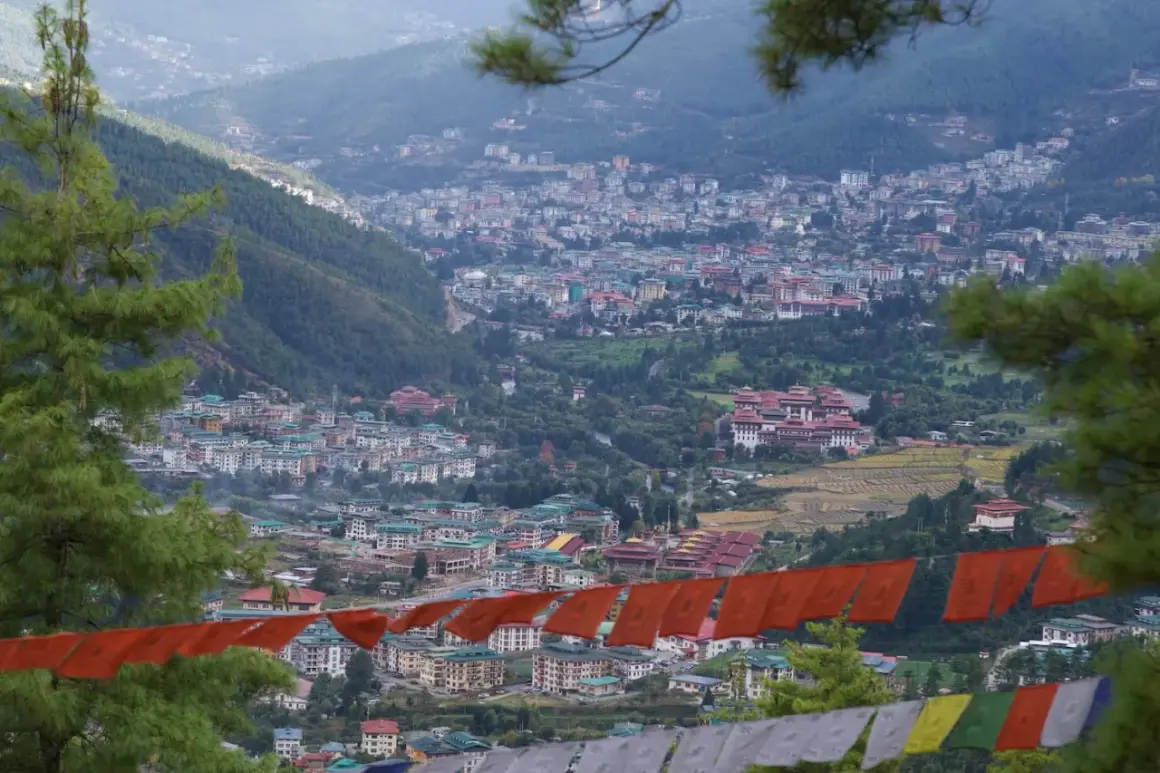 Widok na stolicę Bhutanu, Thimphu, z góry. W oddali widać gęsto zabudowane wzgórza, a na pierwszym planie wiszą kolorowe flagi modlitewne.