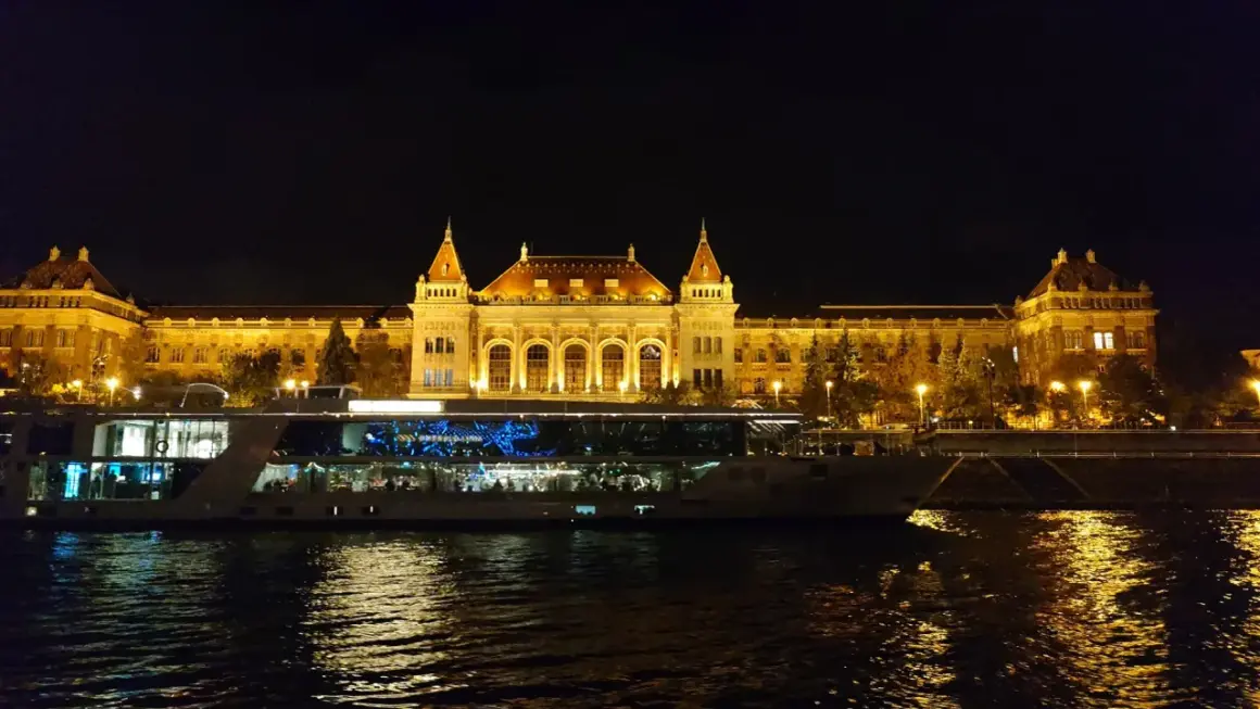 Nocny rejs po Dunaju w Budapeszcie. Oświetlony budynek i statek wycieczkowy odbijają się w wodzie.