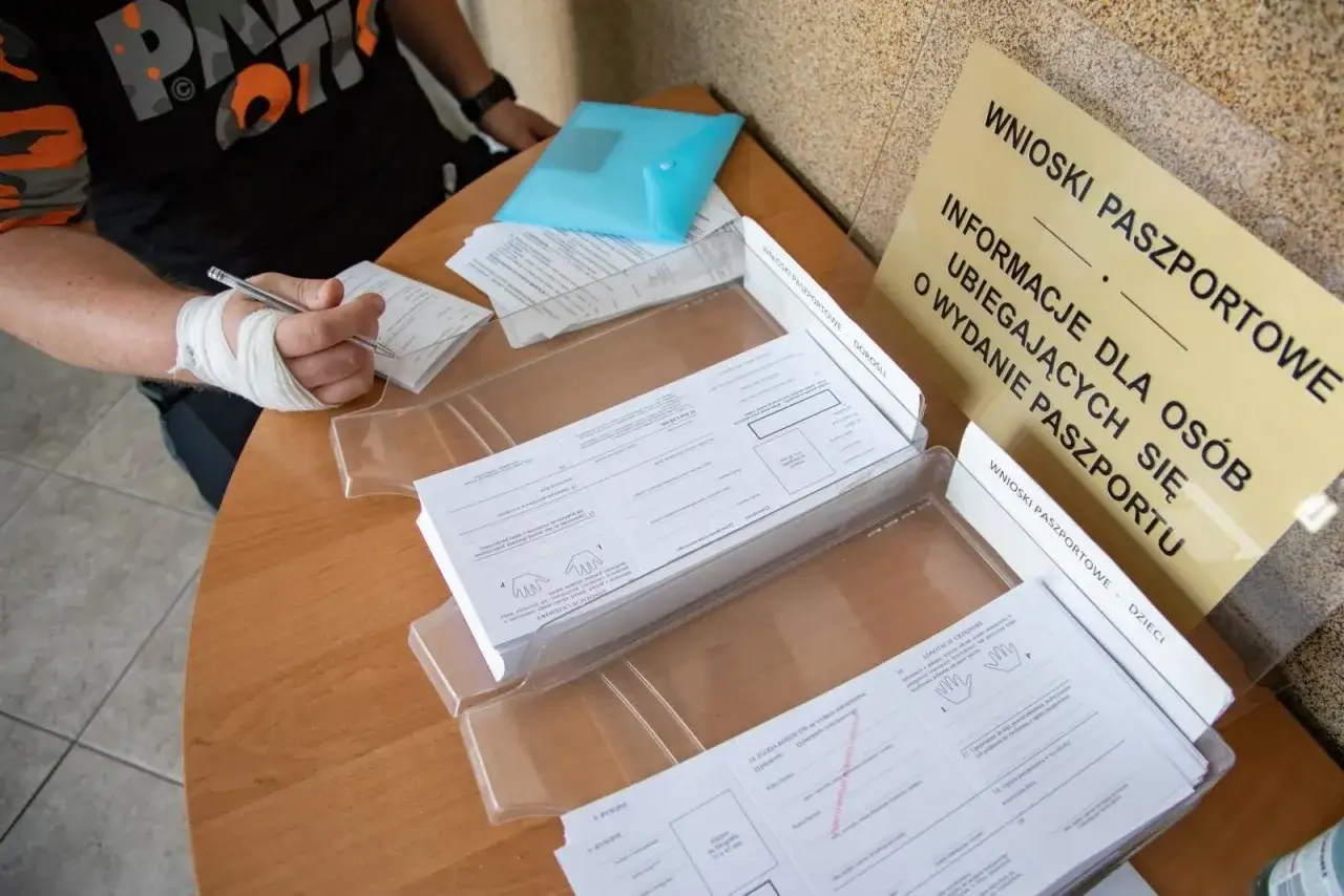 Osoba z zabandażowaną ręką wypełnia wniosek o paszport w Siedlcach. Na stole leżą formularze i tablica informacyjna.