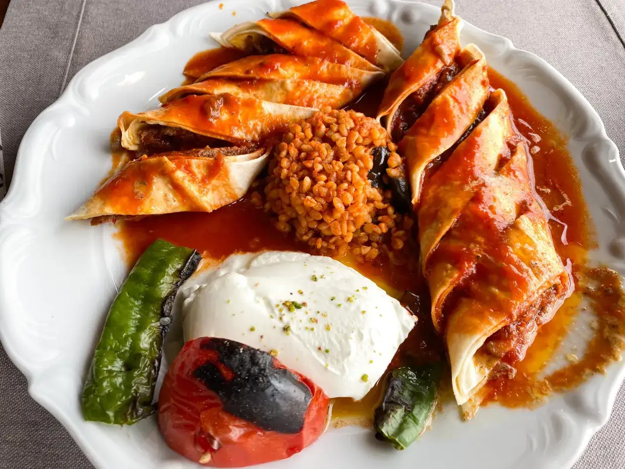 Pyszne tureckie jedzenie: rolady z mięsem w sosie pomidorowym, bulgur, pieczona papryka i pomidor, oraz porcja jogurtu z pistacjami.