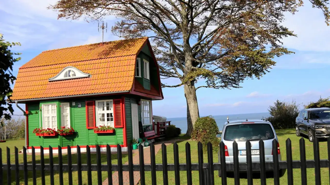 Zielony domek z czerwonym dachem i okiennicami, idealny na wycieczki dla emerytów i rencistów. Obok stoją dwa samochody, a w tle widać morze.