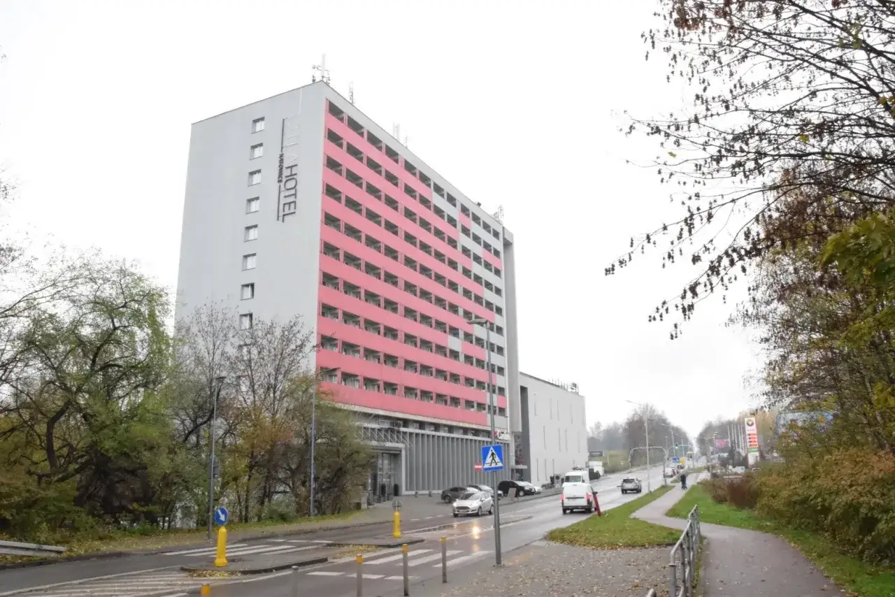 Wysoki budynek hotelu Park Inn Hotel Katowice z różowymi balkonami, otoczony drzewami i ulicą z samochodami.