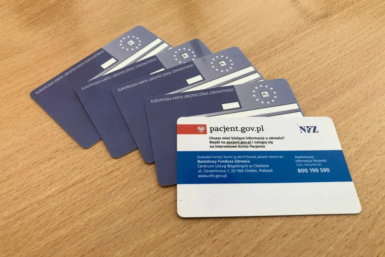 Kilka Europejskich Kart Ubezpieczenia Zdrowotnego (EKUZ) leży na drewnianym blacie. Jedna z kart zawiera informacje o pacjent.gov.pl i numer telefonu NFZ.