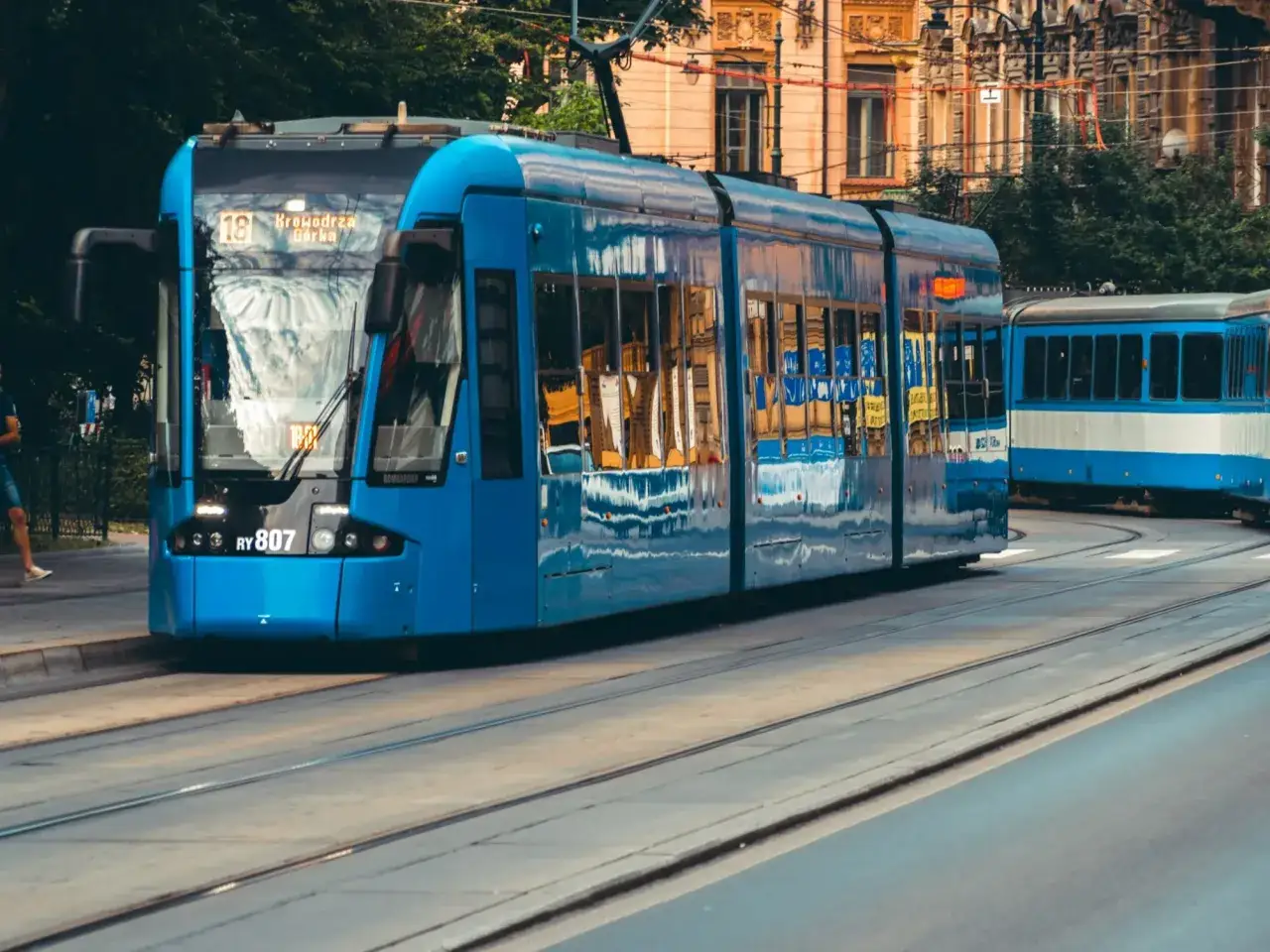 Błękitne tramwaje na torach, idealne na city break z Krakowa. W tle kamienice i drzewa.