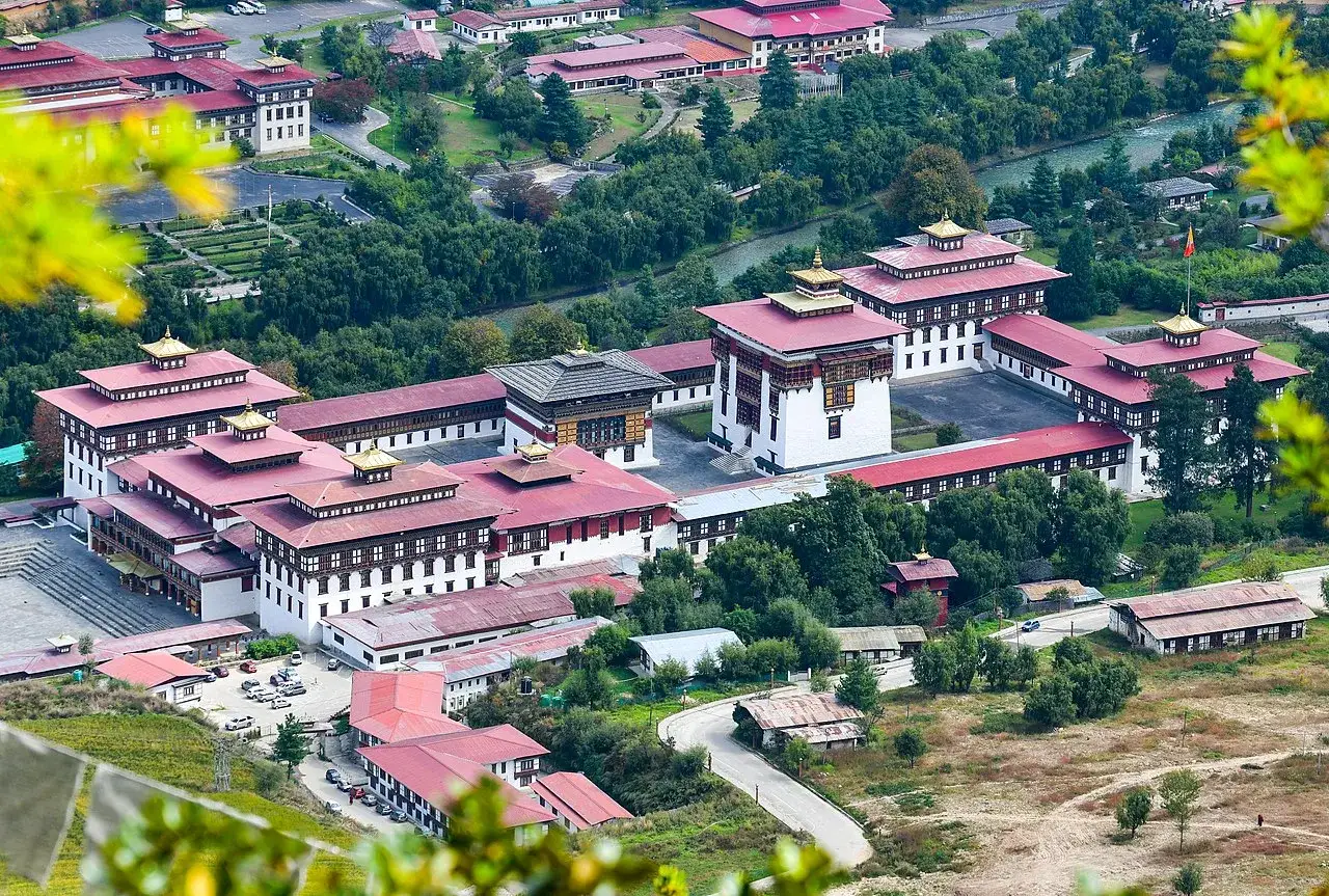 Thimphu, stolica Bhutanu, z widokiem na kompleks budynków z czerwonymi dachami i złotymi zdobieniami, otoczony zielenią.