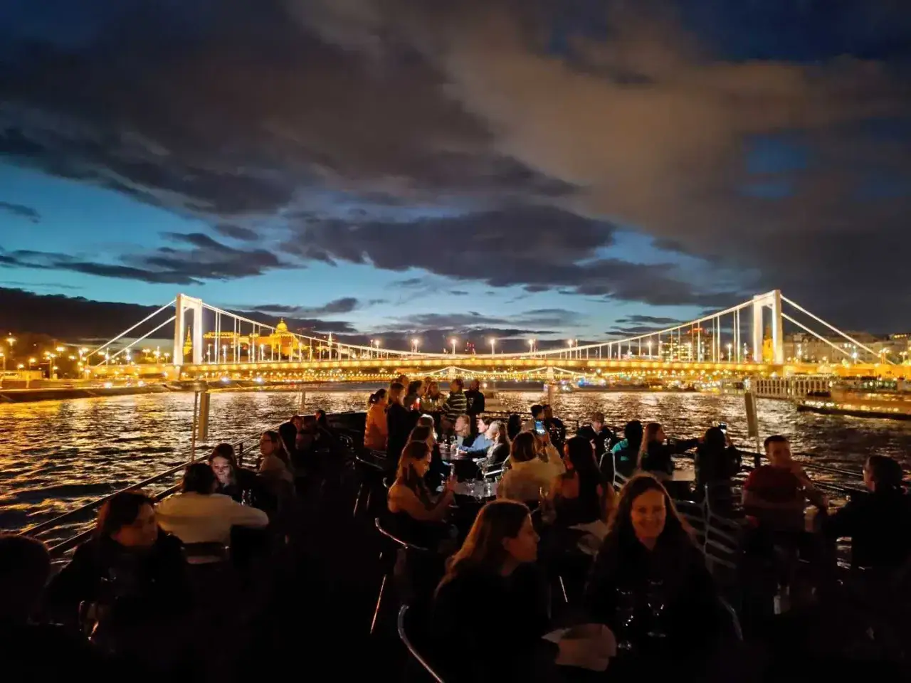 Nocny rejs po Dunaju w Budapeszcie. Ludzie siedzą przy stolikach na pokładzie statku, podziwiając oświetlony Most Elżbiety i panoramę miasta.