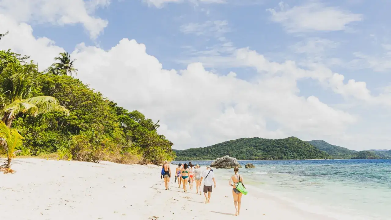 Grupa ludzi spaceruje po piaszczystej plaży na Filipinach, ciesząc się wakacjami. Tropikalna zieleń i turkusowe morze tworzą rajski krajobraz.