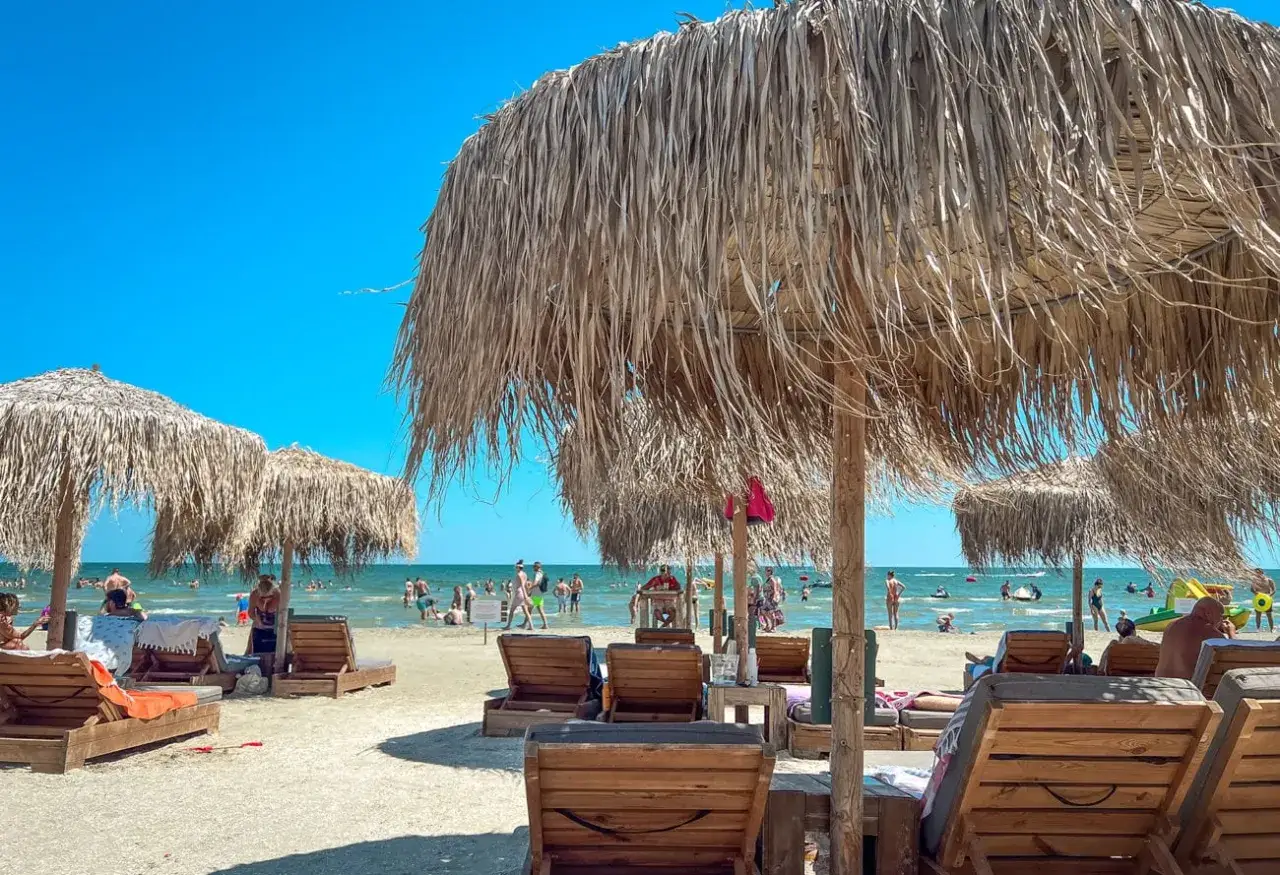 Słoneczne rumuńskie plaże z leżakami i parasolami z trawy. W tle widać ludzi kąpiących się w morzu.