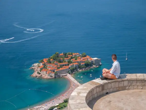 Mężczyzna siedzi na murze, patrząc na wyspę Sveti Stefan. Czy Czarnogóra jest w Unii? Piękny widok na Adriatyk.