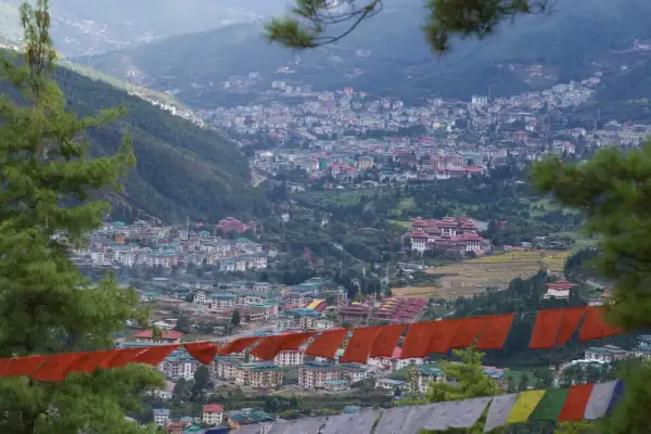 Widok na stolicę Bhutanu, Thimphu, z góry. W oddali widać gęsto zabudowane wzgórza, a na pierwszym planie wiszą kolorowe flagi modlitewne.