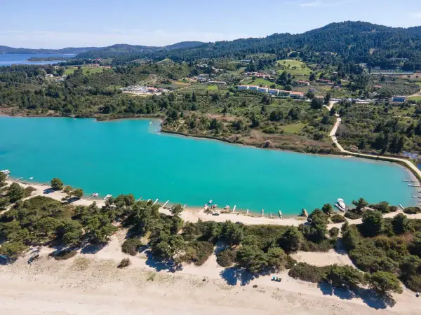 Turkusowa laguna na Kassandrze, otoczona zielonymi wzgórzami i sosnowym lasem. W oddali widać zabudowania i plażę z łodziami.