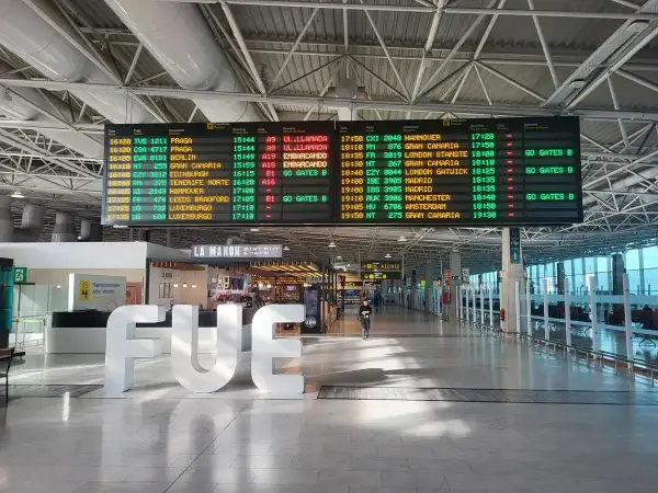 Tablica odlotów na lotnisku, z widocznym napisem "FUE". Informacje o lotach, w tym boarding, wskazują na podróż.